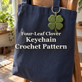 Green four-leaf clover crochet keychain hanging from a navy canvas tote on a wooden chair beside a window, with the product title across the bag.