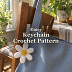 White crochet daisy keychain with a yellow centre hanging from a blue canvas bag on a wooden chair, with the title Daisy Keychain Crochet Pattern.