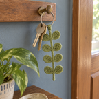 Sage-green crochet eucalyptus keychain and two house keys hanging from one silver ring on a brass hook against a blue hallway wall.
