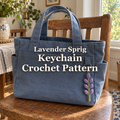 Purple crochet lavender sprig keychain hanging from the front pocket of a blue canvas bag on a wooden chair beside a sunny window.