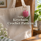 Pink crochet tulip keychain with a green stem and leaf hanging from a canvas tote beside a home window, with the title Tulip Keychain Crochet Pattern.
