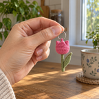 An adult hand holding a small pink crochet tulip keychain by its silver ring beside a kitchen window.