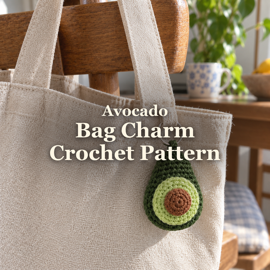 Green crochet avocado charm with pale-green flesh and a brown pit hanging from a linen tote on a wooden chair, with the title Avocado Bag Charm Crochet Pattern.
