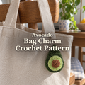 Green crochet avocado charm with pale-green flesh and a brown pit hanging from a linen tote on a wooden chair, with the title Avocado Bag Charm Crochet Pattern.