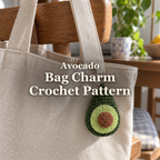 Green crochet avocado charm with pale-green flesh and a brown pit hanging from a linen tote on a wooden chair, with the title Avocado Bag Charm Crochet Pattern.