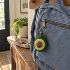 Crochet avocado charm hanging by a silver keyring from the zipper of a blue canvas backpack in a home entryway.
