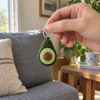 An adult hand holding a small green crochet avocado charm by its silver keyring in front of a linen sofa.