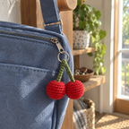 A red crochet cherry pair with green stems hangs from the zipper pull of a blue crossbody bag beside a sunny home window.