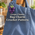Purple crochet grape cluster with a green leaf hanging from a blue canvas tote on a wooden chair beside a sunny window, with the title Grape Cluster Bag Charm Crochet Pattern.