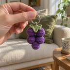 An adult hand holds a small silver keyring with a purple crochet grape cluster and green leaf in front of a linen sofa.