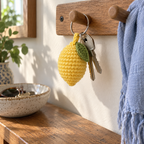 Yellow crocheted lemon charm with one green leaf hanging beside two house keys from a wooden peg above an entryway console.