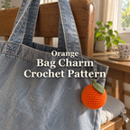 Orange crochet bag charm with a green leaf hanging from a denim tote on a wooden dining chair by a window, with the title Orange Bag Charm Crochet Pattern.