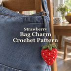 Red crochet strawberry bag charm with a green leafy crown and off-white seeds hanging from a denim bag on a wooden chair, with the title Strawberry Bag Charm Crochet Pattern.