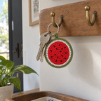 A round red, white and green crochet watermelon charm hangs beside two house keys from a brass hook on a wooden key rail near a front door.