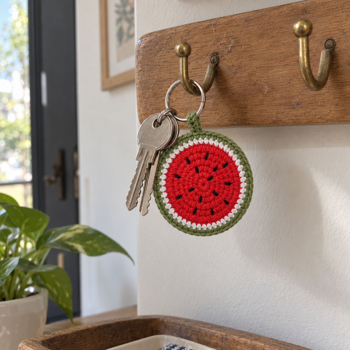 A round red, white and green crochet watermelon charm hangs beside two house keys from a brass hook on a wooden key rail near a front door.