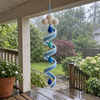 A white cloud crochet wind spinner with a white-edged blue spiral and blue and teal raindrops hangs beneath a wooden porch beam beside a rainy garden.