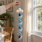 An adult hand holds the blue yarn loop of a white cloud crochet wind spinner, with the full blue spiral and blue and teal raindrops visible beside a home window.