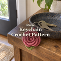 A pink crochet rose keychain with one green leaf hangs from house keys over the rim of a ceramic dish on a wooden table by the door, with the title Rose Keychain Crochet Pattern.