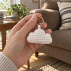 An adult hand holding a white crocheted cloud bag charm by its silver ring in front of a brown sofa.