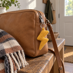 Amber crochet lightning bolt attached to the zipper of a tan leather crossbody bag on a wooden entryway bench beside a folded scarf.