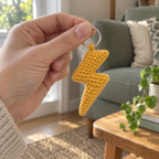 Adult hand holding an amber crochet lightning bolt by its silver ring beside a window, with a sage sofa and wooden side table behind.