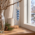 Ice-blue crochet spiral ornament with an off-white snowflake topper hanging from a twig by a window, beside a glass vase and ceramic mug.