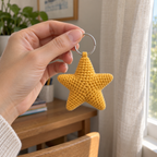 An adult hand holds a small yellow crochet star charm by its silver keyring beside a window.