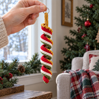 An adult hand holding the silver ring of a red, cream, green and gold crocheted spiral ornament beside a Christmas tree and a window.