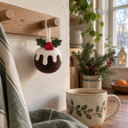 A crochet Christmas pudding ornament hangs by its white loop from a wooden kitchen peg near a ceramic mug and a window.