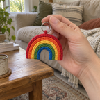 An adult hand holding a small five-colour rainbow crochet charm by its silver keyring in a living room.