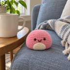 Pink crochet mochi with a smiling brown face and cream front patch sitting on a blue sofa cushion beside a wooden side table and mug.