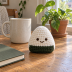 White triangular crochet onigiri with a green lower band and smiling brown face standing on a wooden desk beside a mug, notebook and houseplant.