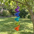 A purple, blue, teal, green and orange crochet wind spinner with matching balls hangs from a tree branch over a sunny lawn.