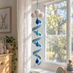 A sky-blue crochet wind spinner with a white cloud and blue and teal raindrops hangs beside a sheer curtain in a bright nursery window.