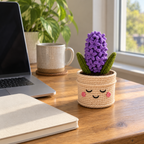 A purple crochet hyacinth with two green leaves stands in a cream smiling pot on a wooden desk beside a laptop, notebook and mug.