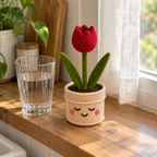 A red crochet tulip with an open top and two green leaves stands in a cream smiling pot on a wooden kitchen windowsill beside a glass of water.