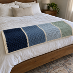 Navy, mid blue, soft blue and mint crochet throw with narrow cream stripes and a cream border folded at the foot of a bed with white linen.