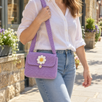 Lilac crochet shoulder bag with a rounded flap, white eight-petal daisy, yellow centre and small button, worn on a sunny street.