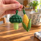 Close view of a bumpy crochet broccoli crown and a yellow corn cob with kernel bobbles and two green husk leaves, beside an adult hand and a mug.
