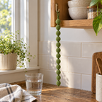 A String of Pearls crochet hanger with small sage-green yarn balls hanging from a kitchen shelf above a wooden counter and water glass.