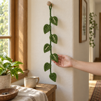 An adult hand gently supports a cream-streaked leaf on a hanging green Pothos crochet vine beside a window.