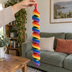 An adult hand holding a rainbow crochet ribbon spinner by its loop in a living room