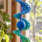 A close view of blue and teal crochet shell lace and a blue ball on a purple cord beside a sunny window