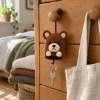 A brown bear crochet key pouch hangs from a wooden drawer knob beside a linen tote, with a silver keyring and key below its open bottom.
