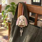 A cream bunny crochet key pouch with pink-lined floppy ears hangs against an olive canvas bag on a wooden chair, with a silver keyring and key below.
