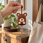 An adult hand holds the brown stopper ball of a small bear crochet key pouch, with its single cord, open bottom, silver keyring and key visible.