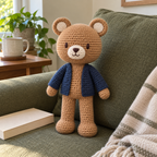 Maple Bear, a honey-brown crochet bear with a cream muzzle and navy cardigan, stands against a green sofa cushion beside a closed book.