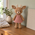 Hazel Deer, a caramel crochet deer with cream antlers and a dusty-rose pinafore, stands on a wooden dresser beside a vase and folded floral shirt.