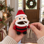 An adult holds a Santa crochet key pouch while the other hand pulls its silver key ring, with two keys below the white rolled mouth and the white ball resting on the red hat tip.