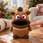 A fingertip touches the thick dark brown rolled lip of a Reindeer crochet key pouch, showing its cotton stitches, tan antlers, red nose and two white eyes.